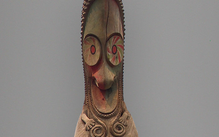 A hollow, wooden Oceanic sculpture with a bird-like face, large round eyes, sharp down-turned beak, and pointed head