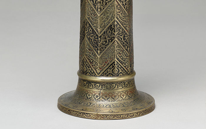 A brass candle stick engraved wtih a zigzag pattern and decorated in inlaid black and red pigment