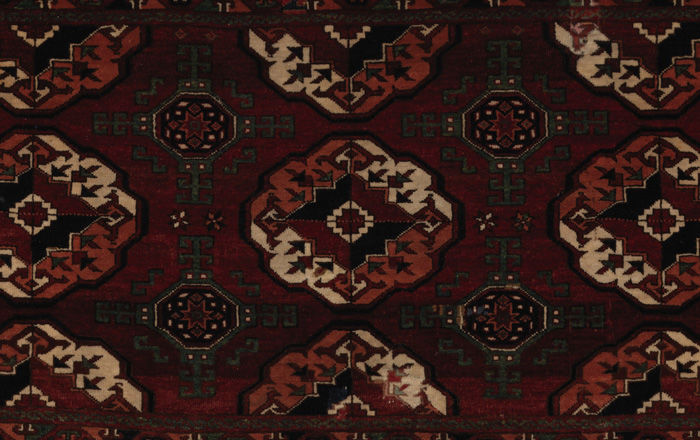 A close-up of a dark brown, burgundy, olive-green and white carpet decorated in a repeating pattern of geometric motifs