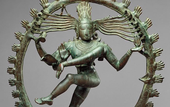 An oxidized copper sculpture of an Indian deity with four arms, standing on one leg dancing, encircled by a ring of stylized fire