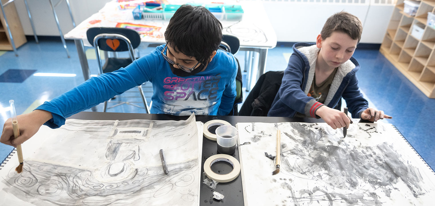Two students sitting at a table while sketching and painting.