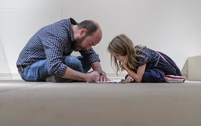 A young girl sits watching a bearded man sketch in the middle of the gallery floor