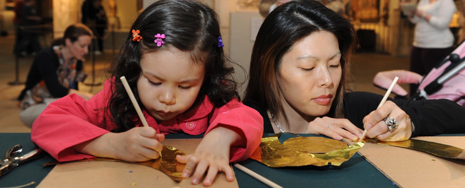 A young girl in a pink shirt and her mother are creating a craft out of gold foil in the middle of the Medieval galleries