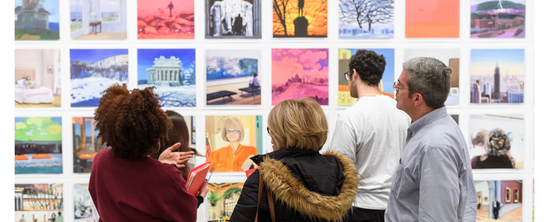 A group of adults stand with backs facing the camera as they observe a work of art on the wall.