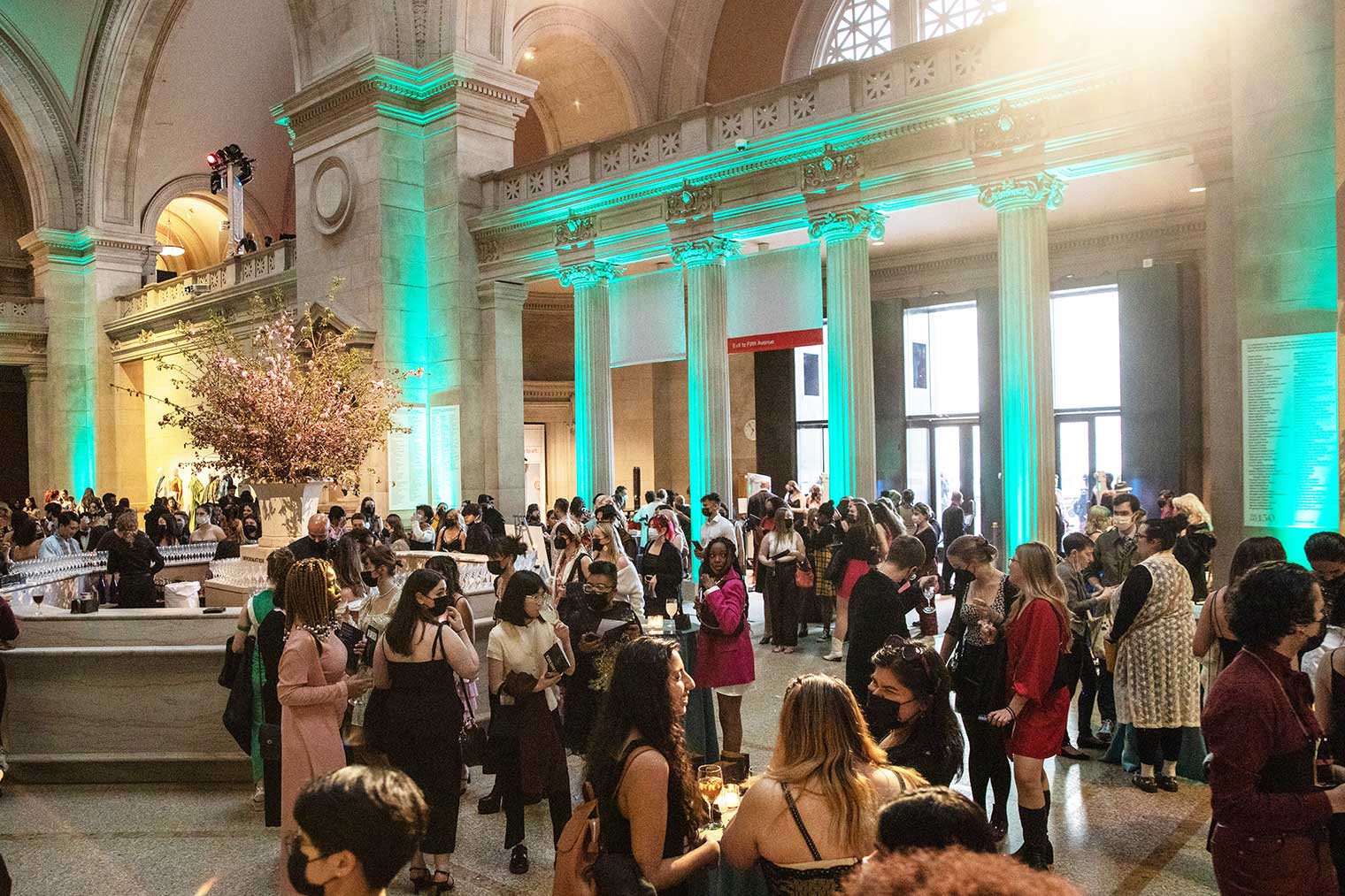 Young people dressed for an evening out gather in a gallery space with very high ceilings and party lights.