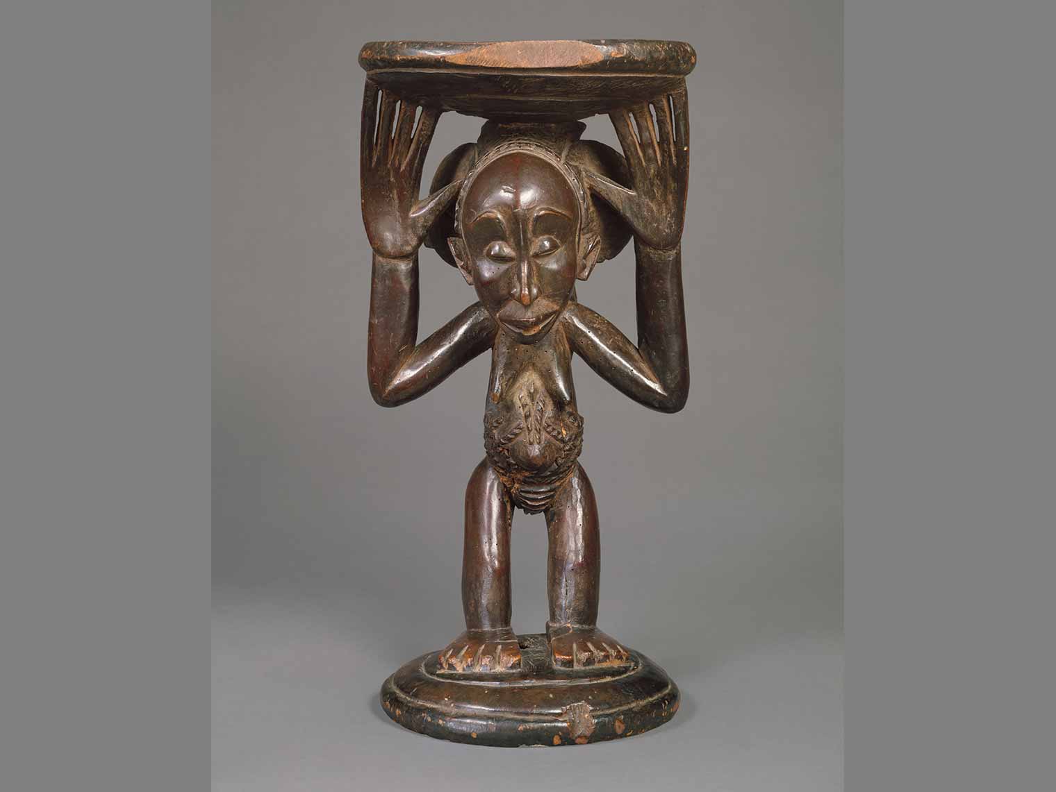 A brown, wooden female body holding the seat of a stool.