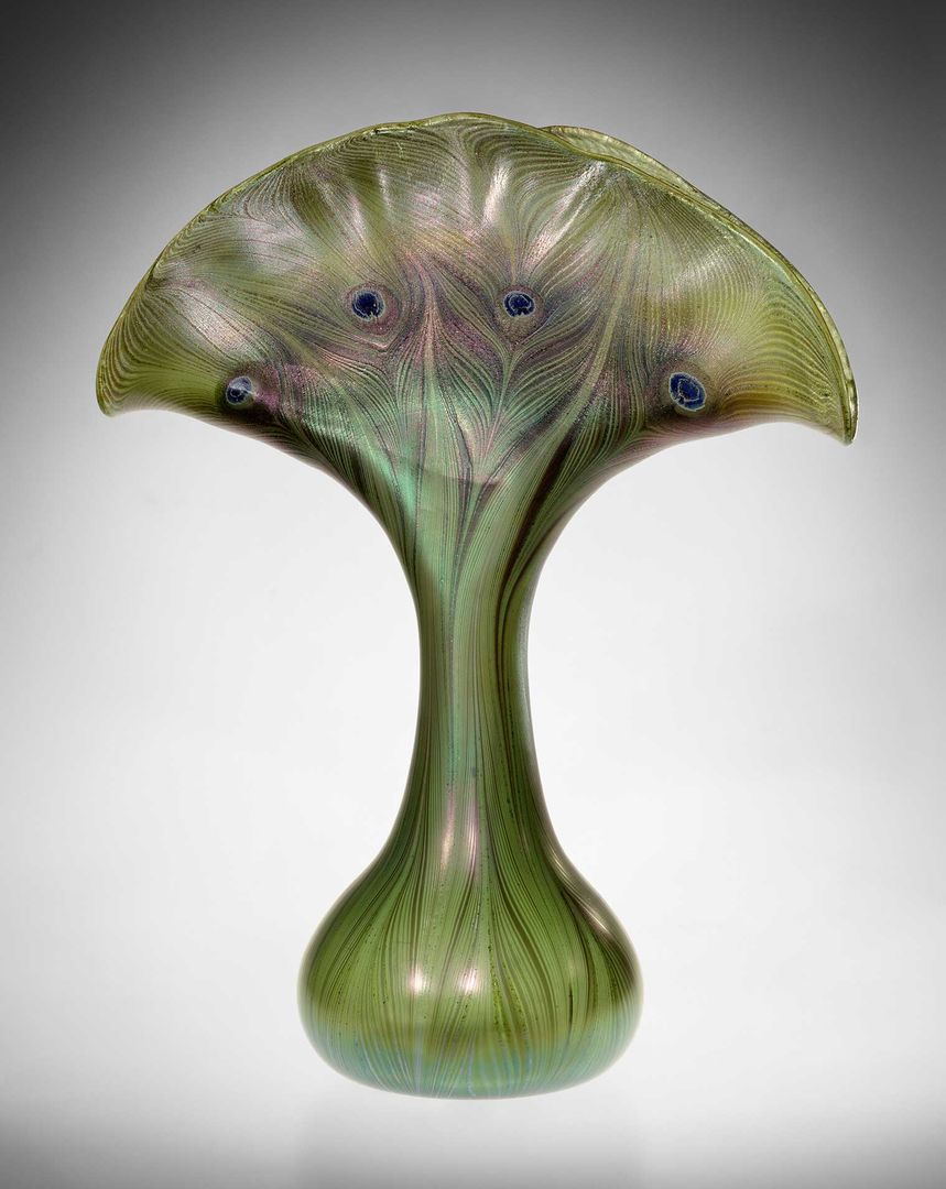 A decorative, green glass vase shaped like a fan at the top and rounded at the base, with peacock feathers designed into the fanned top.