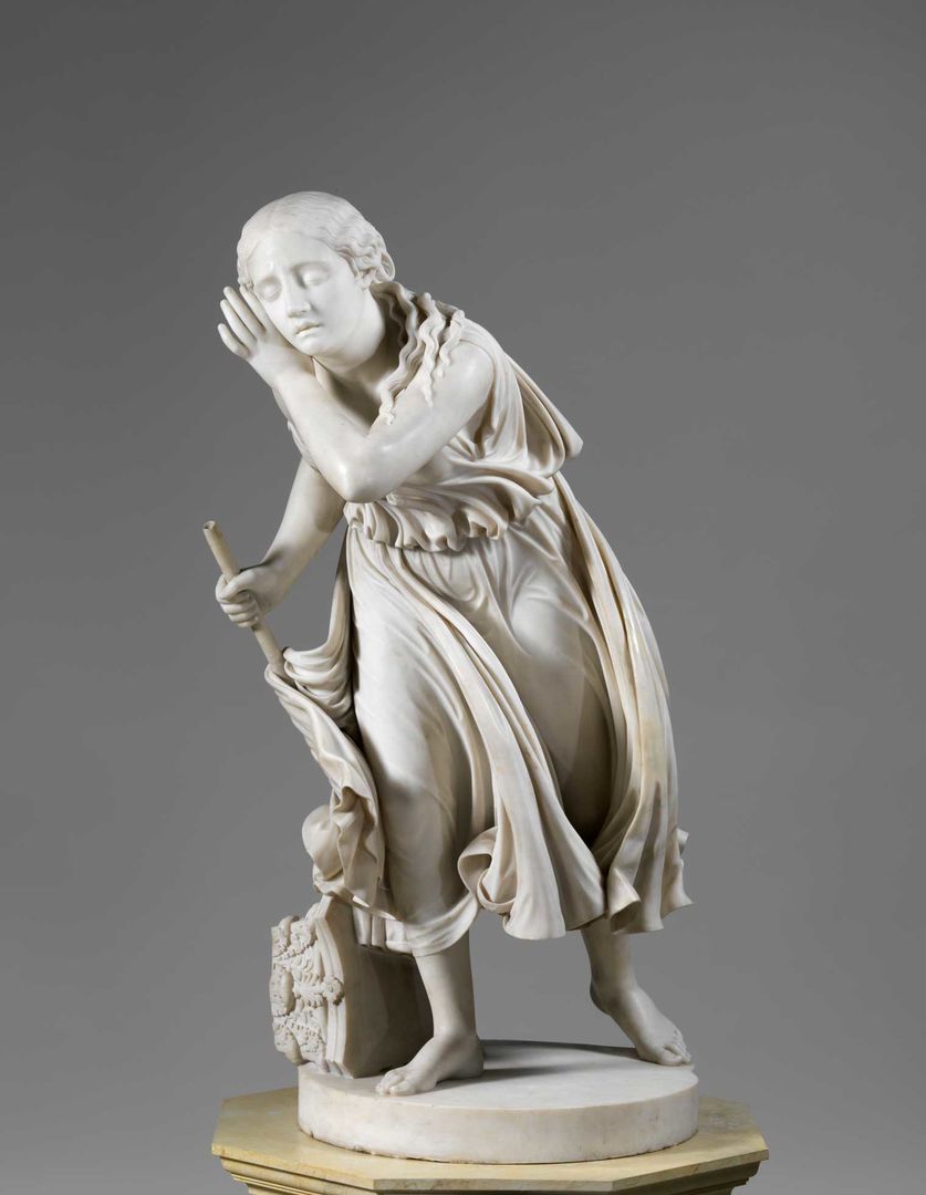 A white marble statue of a girl wearing a cape, holding one hand near her ear and carrying a stick in the other hand