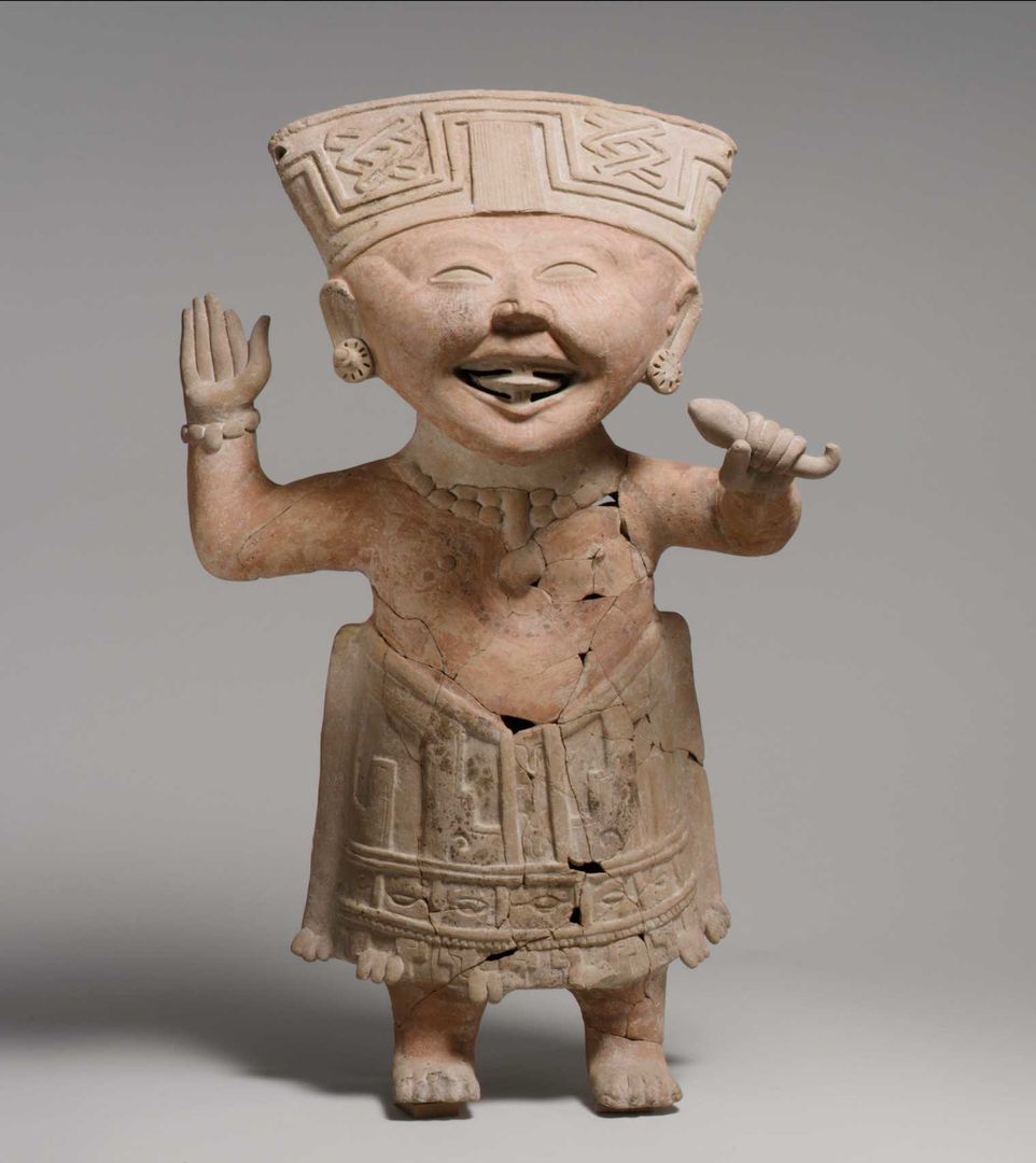 A smiling ceramic figure, nude from the waist up, raising his right hand and holding a rattle in his left hand