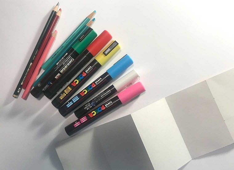 A photo of colored pencils and markers beside the blank folded paper strip
