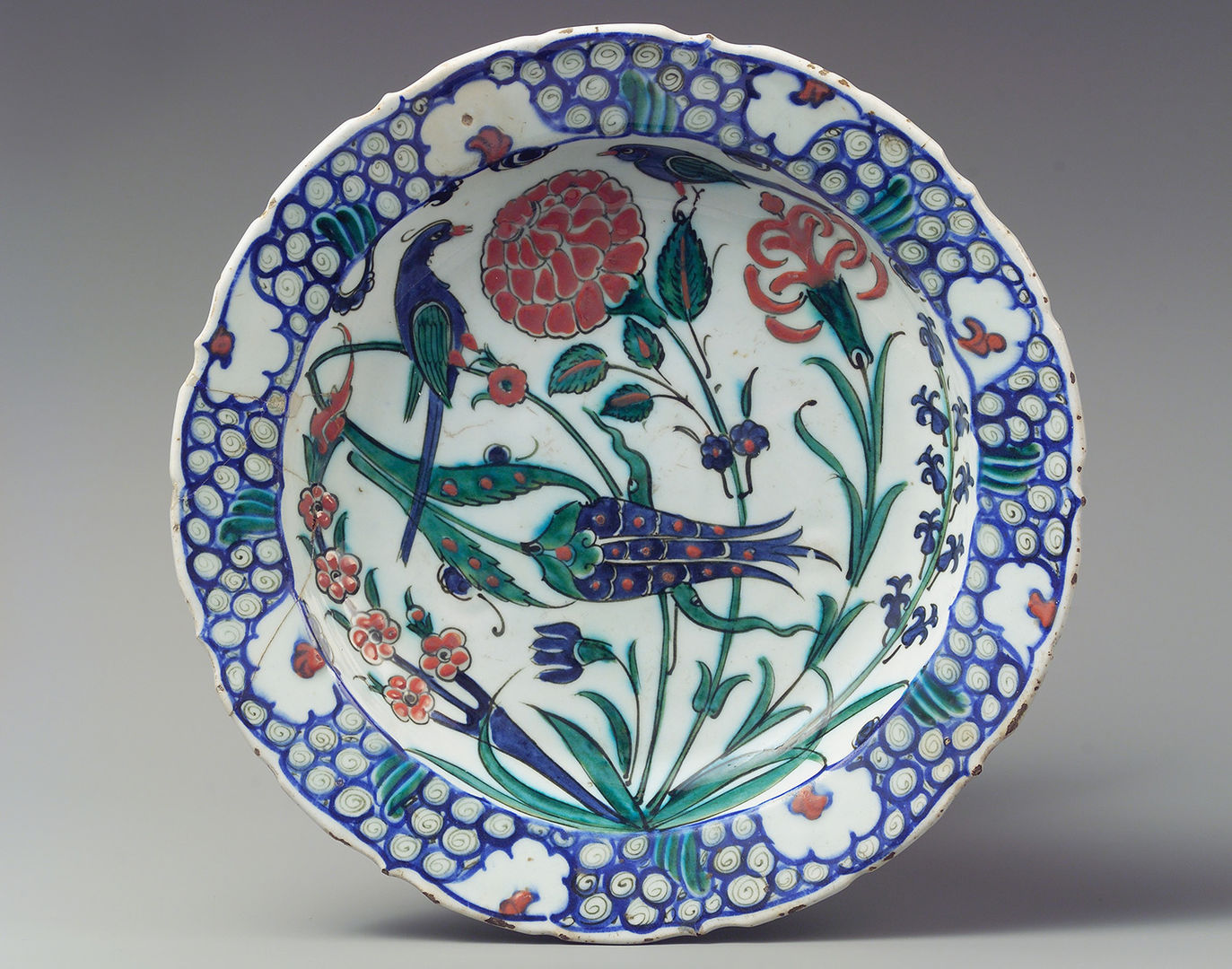 An upright round ceramic plate painted with images of flowers, some bent with red rose buds and some leaning in from the side with a purple tulip head.