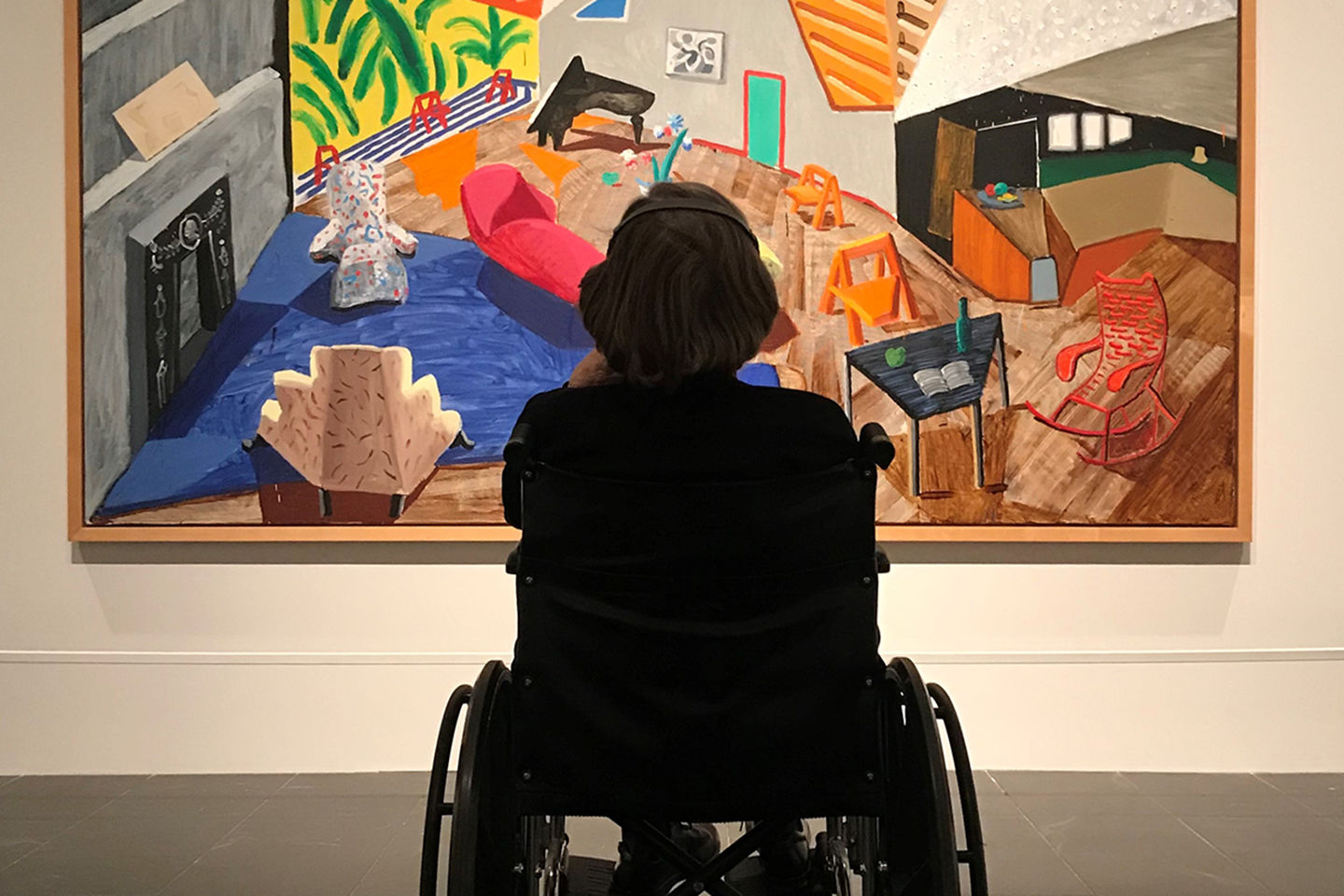 Shown from behind, a visitor sits in a wheelchair looking at David Hockney’s colorful painting, Large Interior, Los Angeles. 