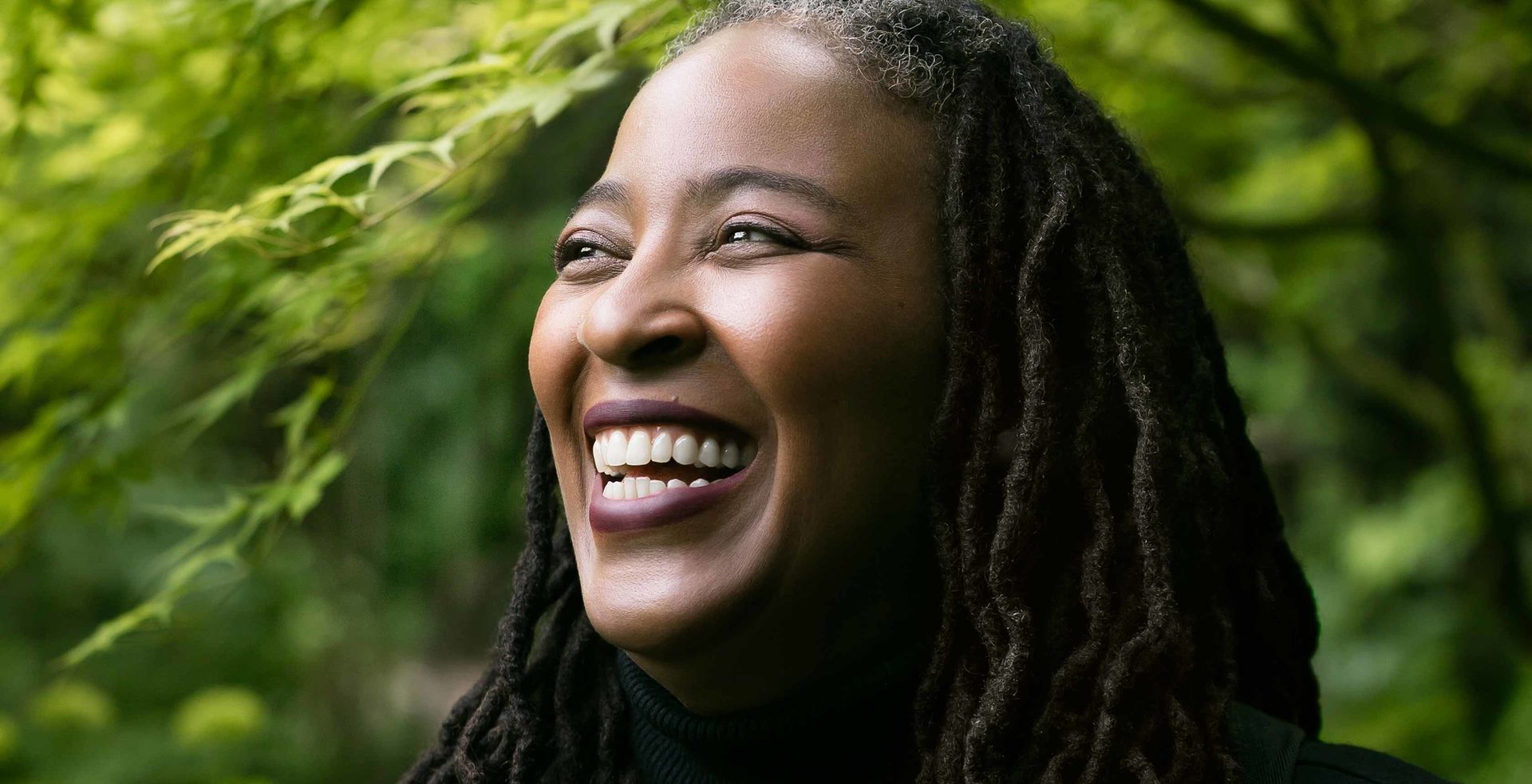 Poet Camille Dungy, a Black woman with long dreadlocks wearing a black turtleneck, laughing and standing among trees