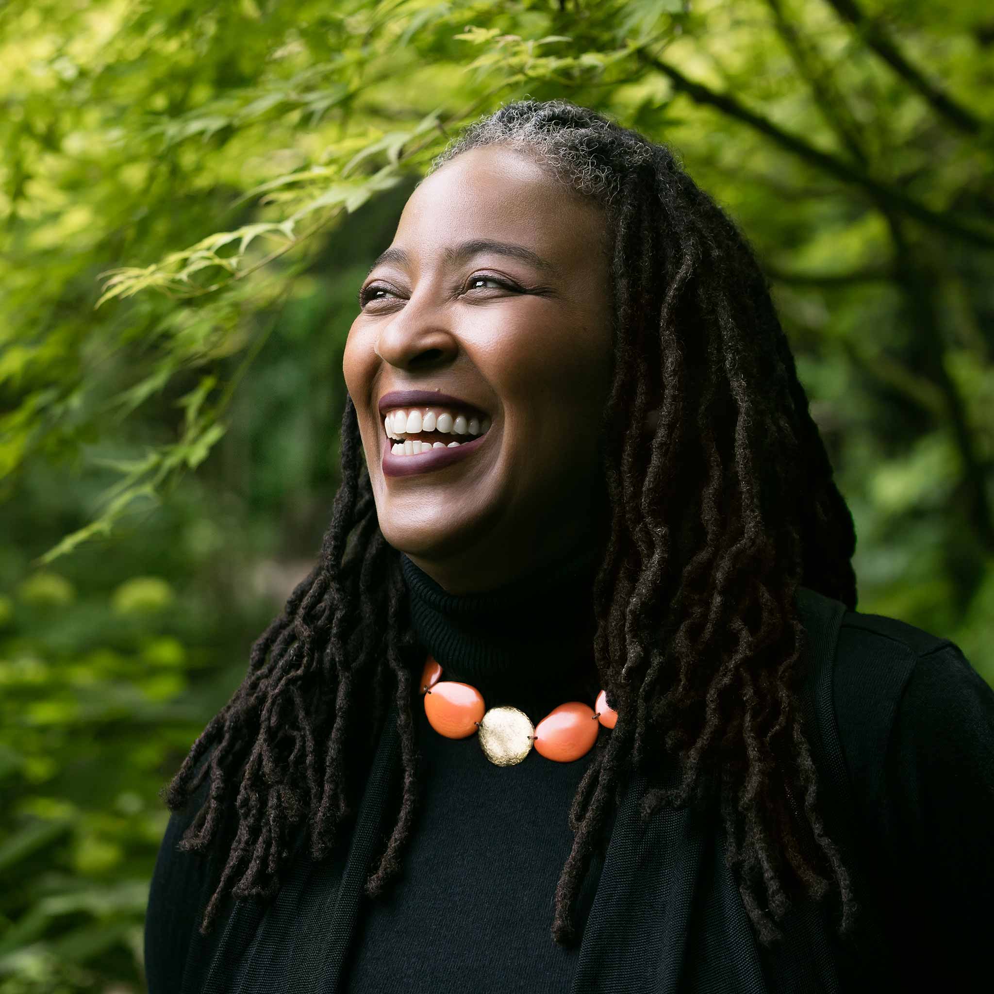 Poet Camille Dungy, a Black woman with long dreadlocks wearing a black turtleneck and a bold orange and gold necklace, laughing and standing among trees