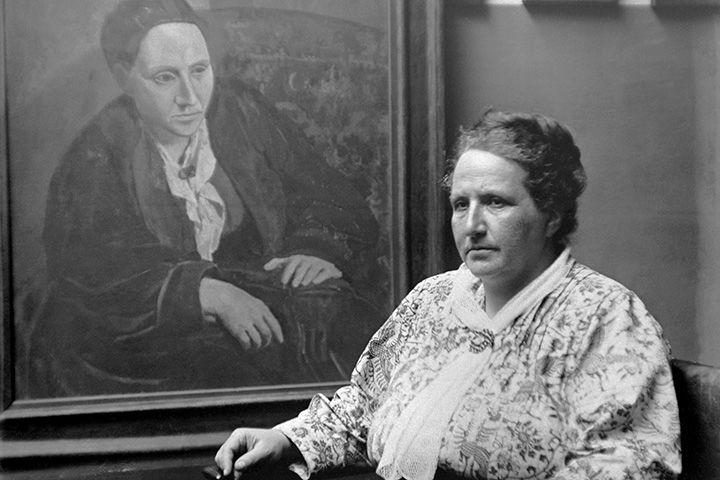 Black and white photograph of a woman wearing a white floral pattern outfit on the right with a potrait in the background done by Picasso of her.