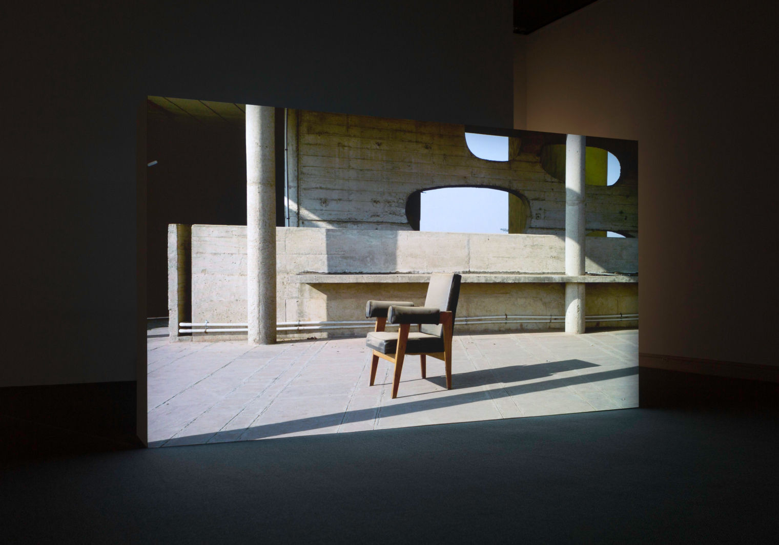 Gallery view of a screen showing a teak chair with gray cushions and armrests in an empty urban setting. 