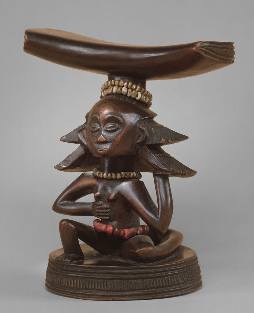 Wood neck rest sculpture of a nude female figure in an asymmetrical pose: the left knee bent up and the right elbow bent downward. Beads are around the figure's head, neck and waist
