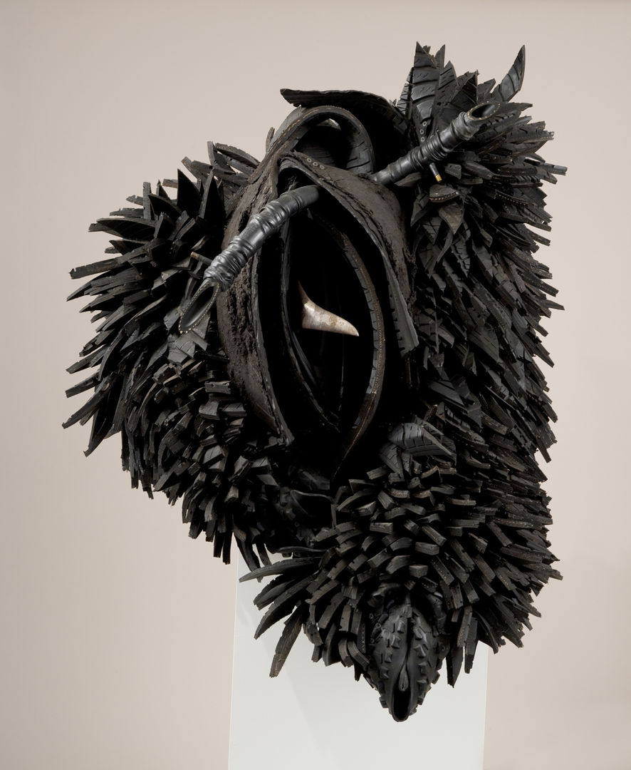 A mound of sliced and gathered tire pieces takes on the look of feathers or scales covering an ambiguous hulking creature. Nestled in this mass are rows of curved rubber pieces that resemble a vulva, from which a sharp piece of steel protrudes. 