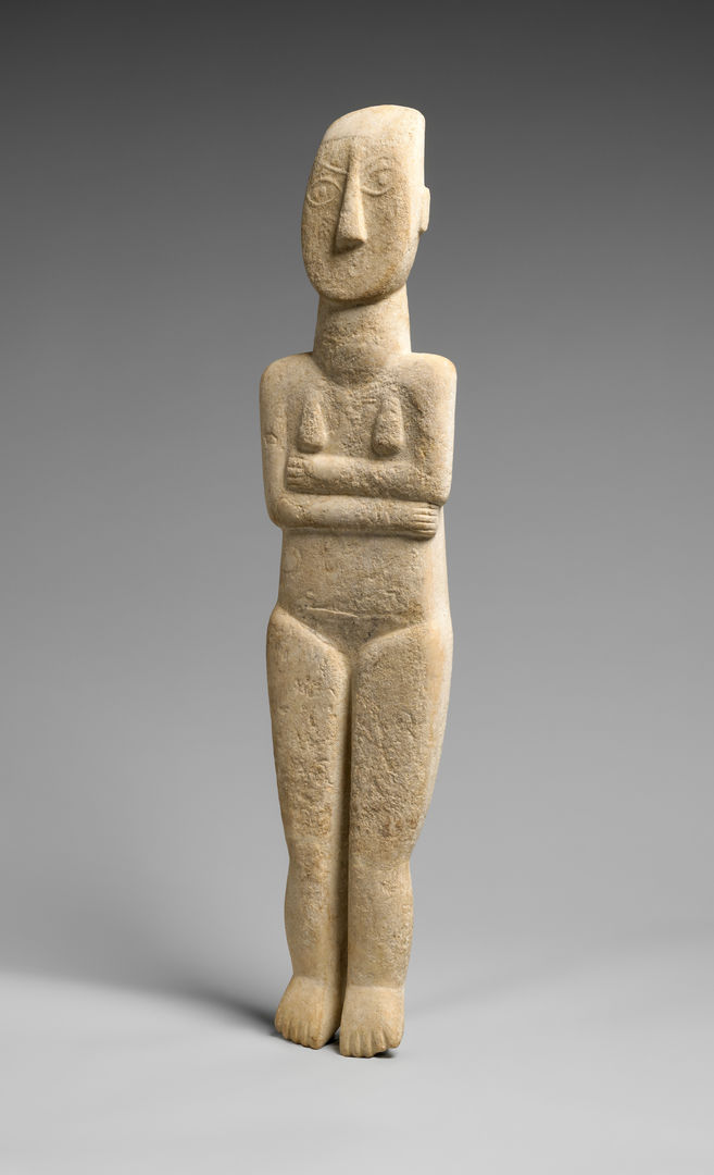 Nude stone Cycladic figure with it's arms crossed. The figure is slightly flat and there are round eyes carved it. It is against a grey background. 