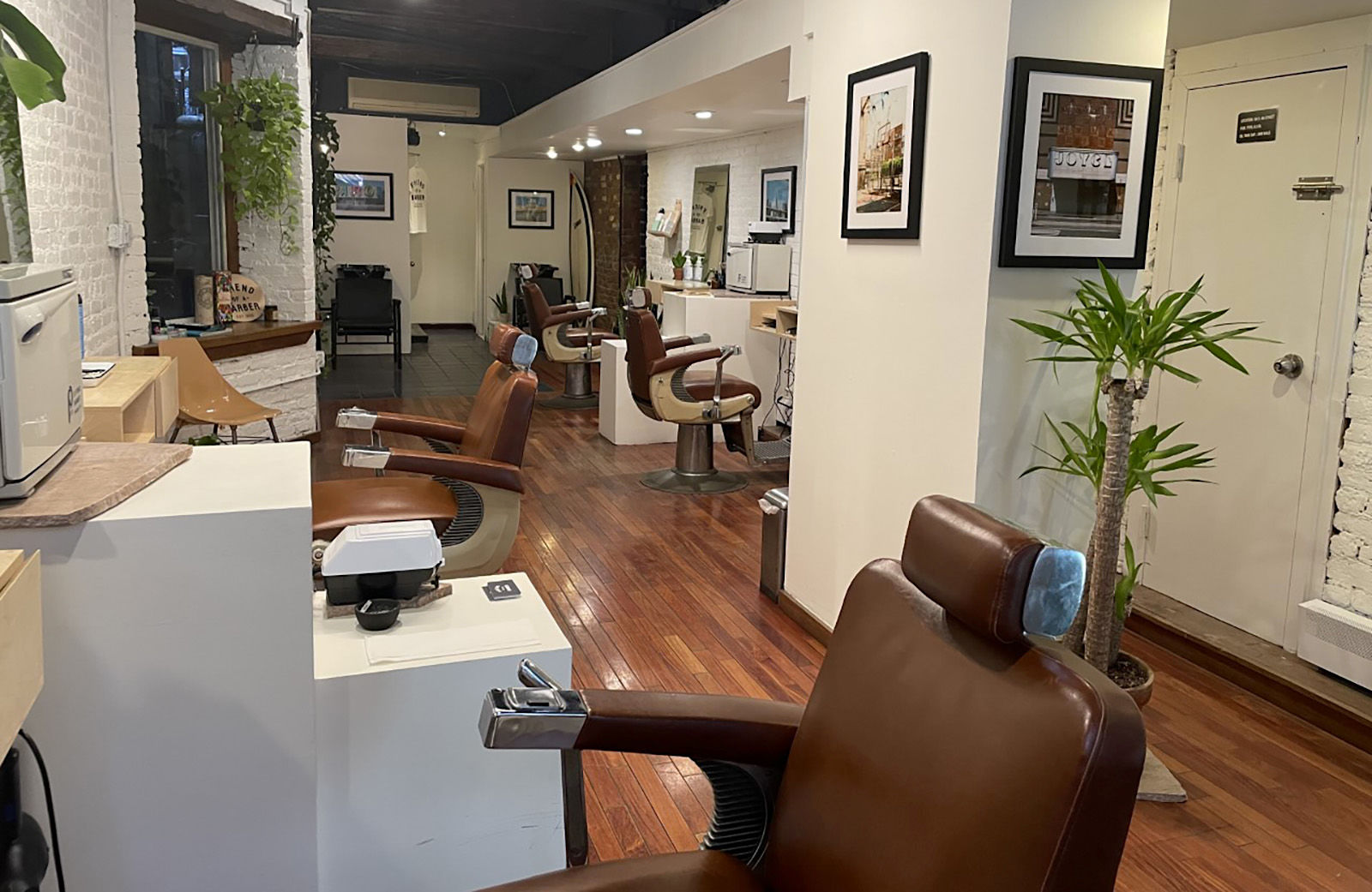 Photograph of the interior of the barbershop showcasing brown leather chairs, plants, and white brick walls.