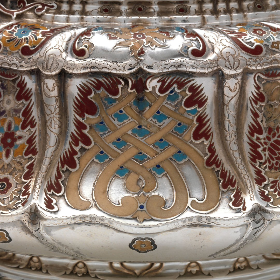 A close up detail of a silver teapot adorned with a symmetrical design of abstract floral motifs rendered in maroon, orange, taupe, and turquoise enamel.