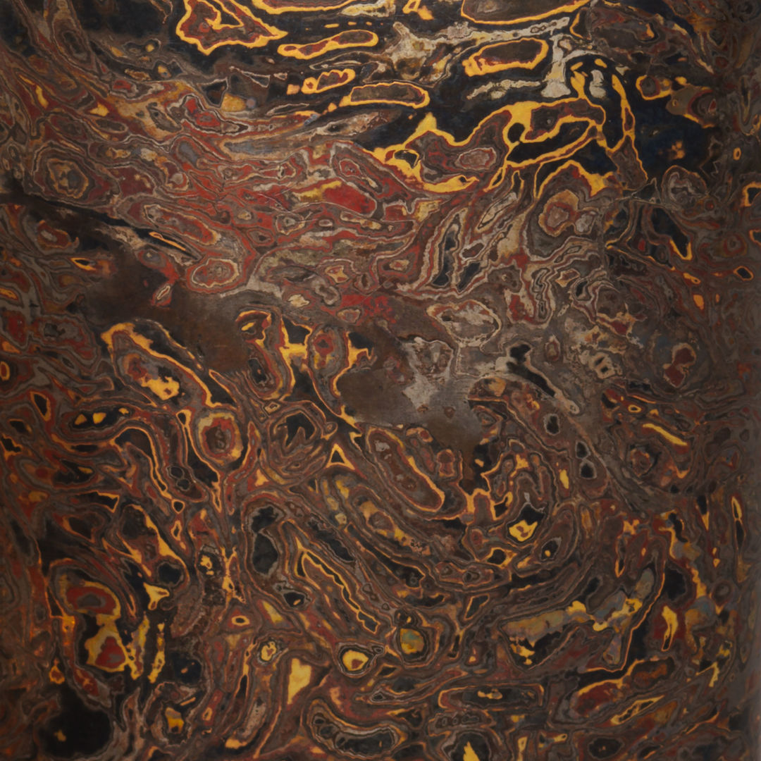 A close up detail of a metal case rendered in a wood grain pattern of deep red, black, brown, and gold metals. 