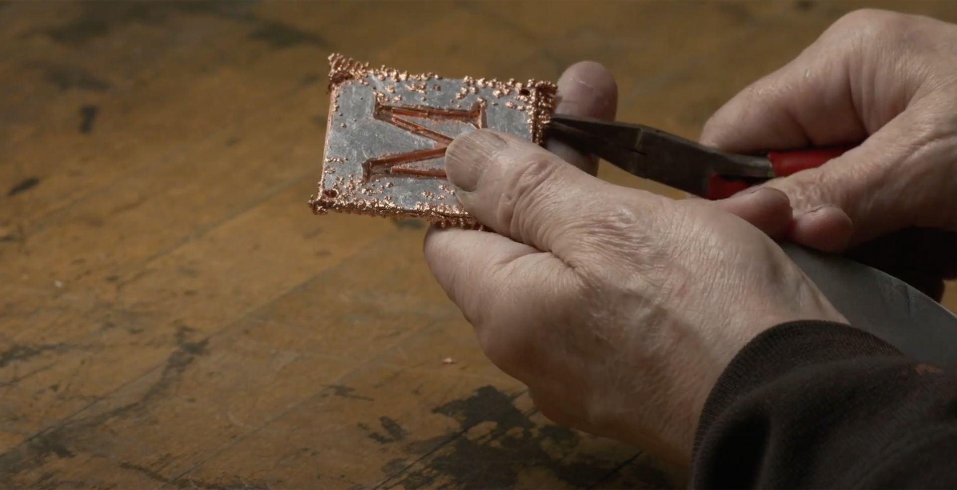  An artisan uses pliers to clip off dendrites of excess copper branching off the surface of a small square silver plate with a copper-filled letter M design in the center.