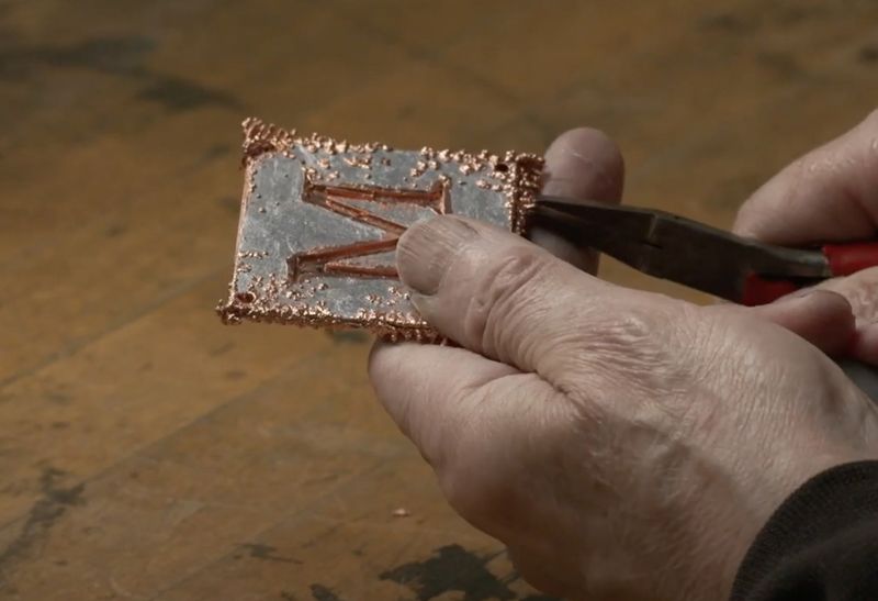 An artisan uses pliers to clip off dendrites of excess copper branching off the surface of a small square silver plate with a copper-filled letter M design in the center.