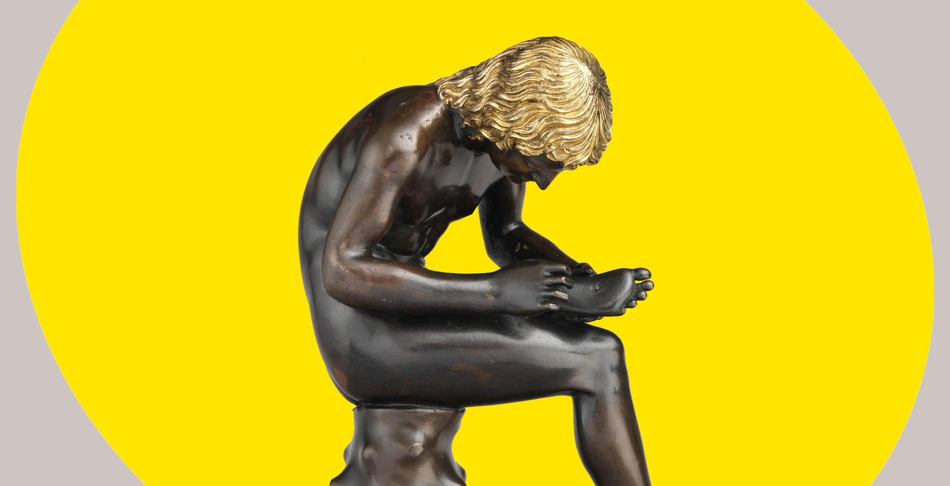 A small bronze sculpture of a seated boy pulling a thorn from the bottom of his foot appears set against a bright yellow ovular spotlight shape in the background.