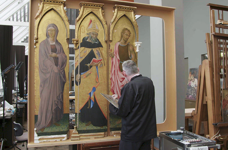 A wide shot of a paintings conservator working on three panels from an Italian altarpiece depicting three religious figures on a gold background. 