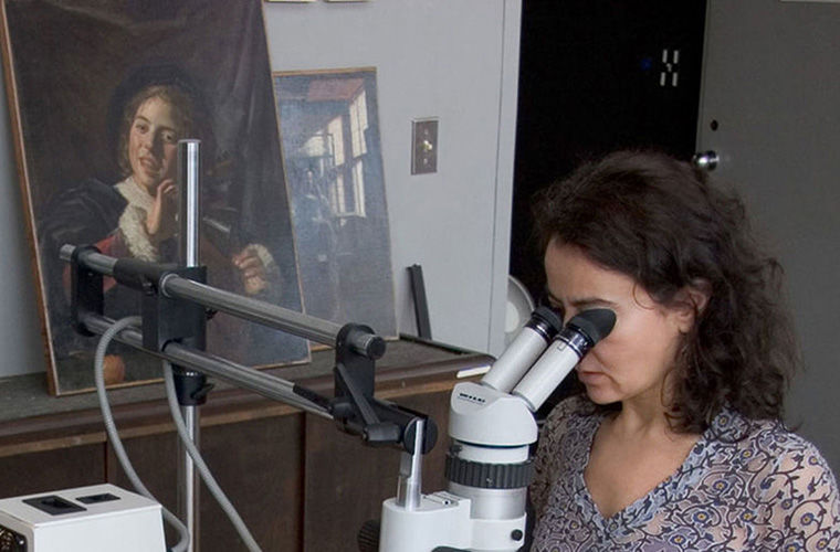 Image of a woman looking at a microscope in a paintings lab with several paintings behind her. 