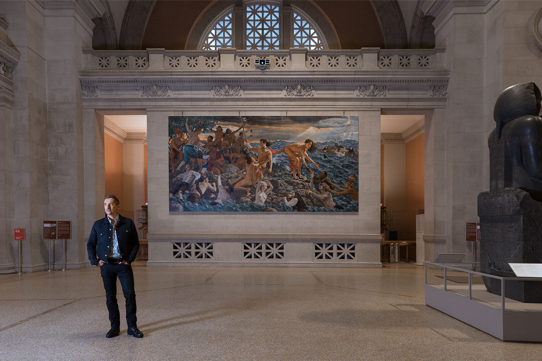 Artist Kent Monkman stands in front of a hall with a colorful painting behind. 