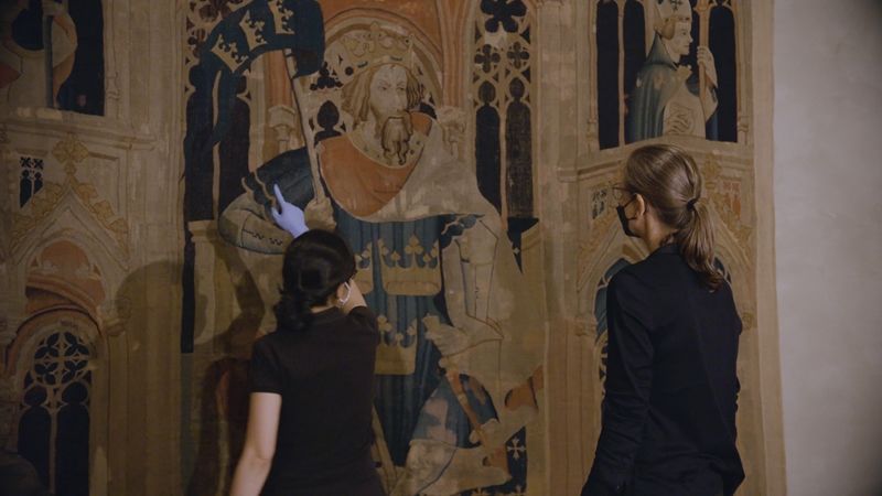 Photo of two women looking at a brown, blue, and red tapestry of a King, woman on left is pointing at the tapestry.