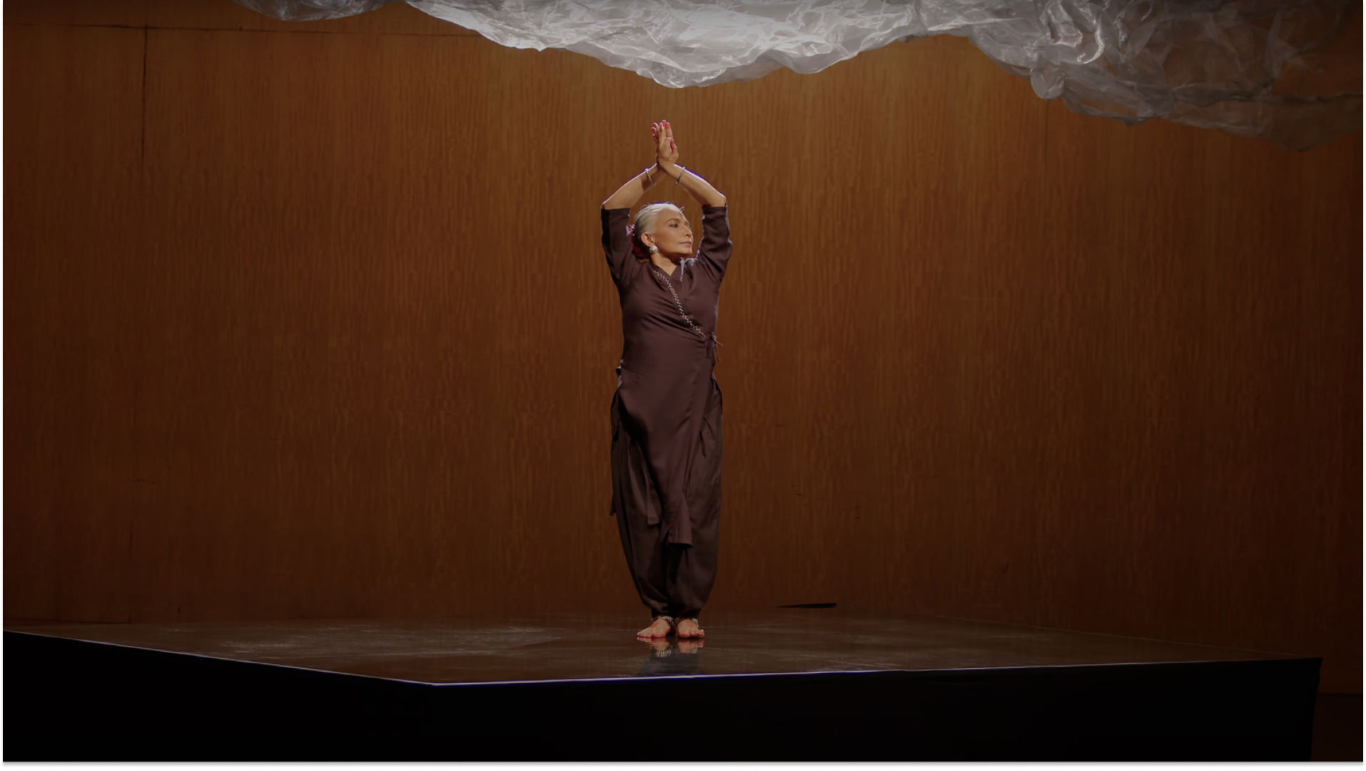 A woman stands on stage wearing a matching brown tunic and pants. Her arms are raised over her head with her palms pressed together. 