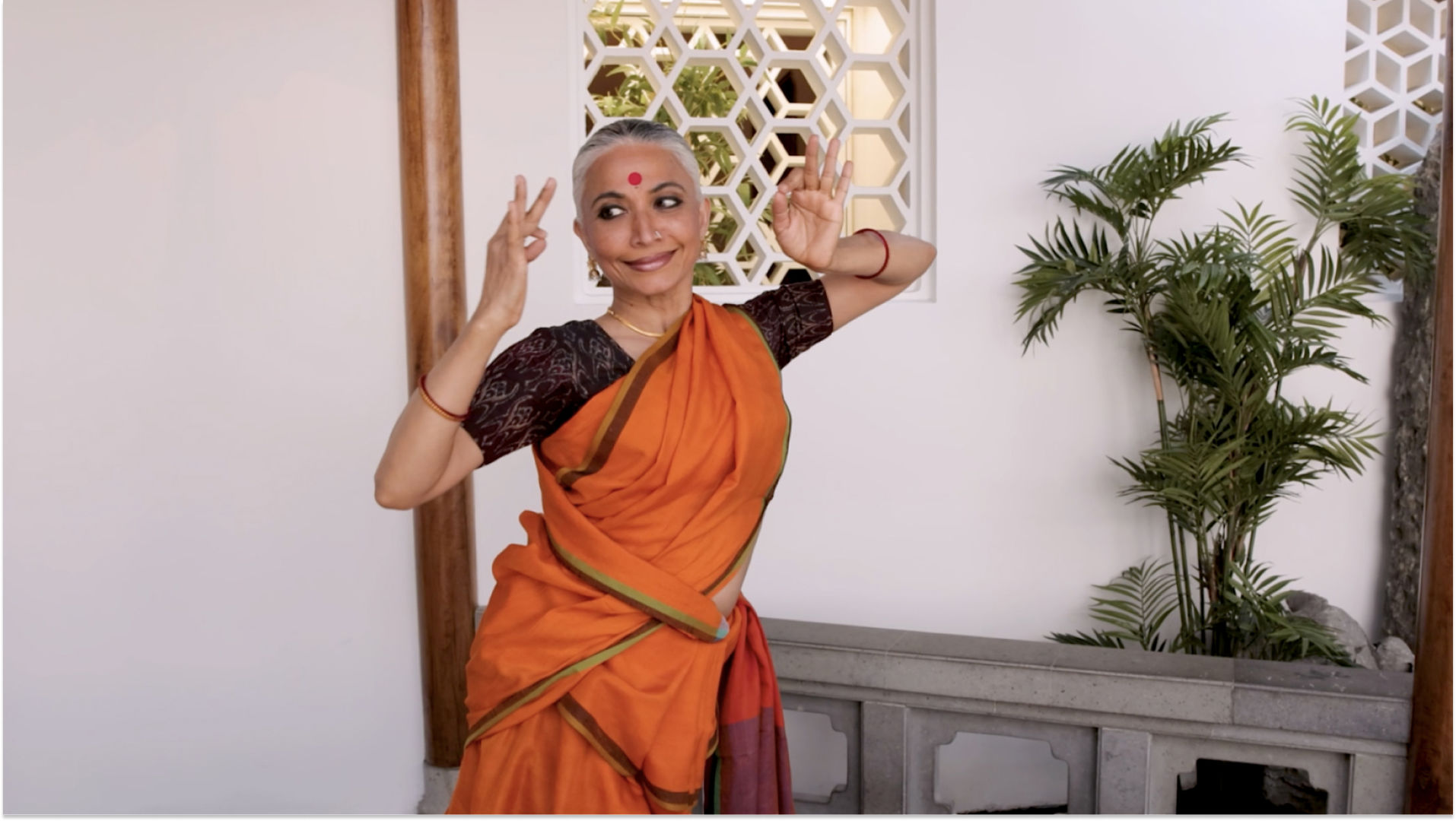 A woman stands wearing an orange sari and red bindi between her eyes. Her arms are held up near her head with her hands making a gesture. Behind her is a whitewall and green plant.