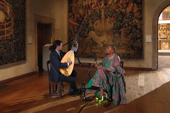 Cécile McLorin Salvant, seated, sings while Dušan Balarin plays a theorbo. They are performing at The Met Cloisters in a room with hanging Medieval tapestries.