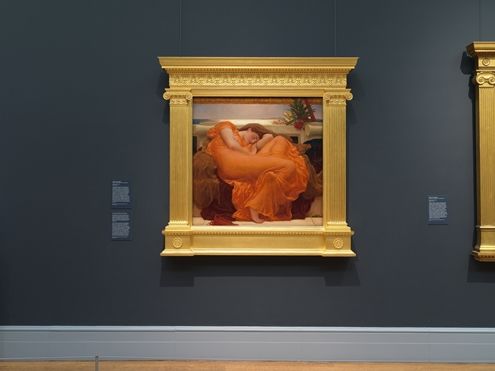 A painting on a wall with a large Greek-inspired frame. The painting is of a woman resting, draped in a bright orange dress.