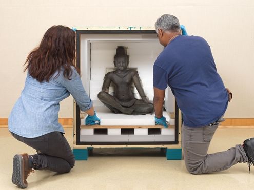 A seated sculpture located inside a crate with two art handlers adjusting the interior of the box