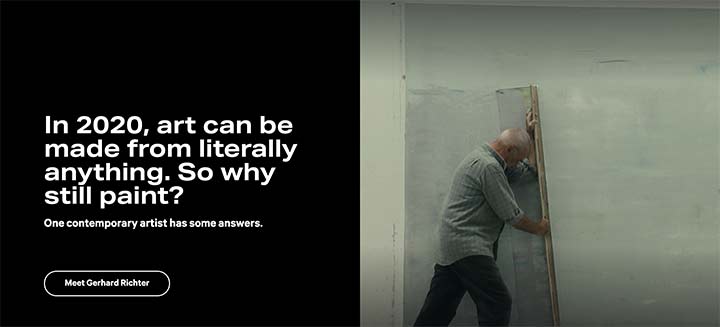 A rectangular graphic image where the left-hand is a simple black background with the following burned in text: "In 2020, art can be made from literally anything. So why still paint? One contemporary artist has some answers. Meet Gerhard Richter." The right-hand side is an image of the artist Gerhard Richter pushes a large vertical squeegee across an oversized green-tinted off-white canvas to reveal gray off-white paint layers underneath. 