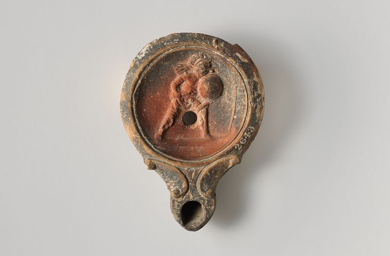 Terracota oil lamp showing a gladiator, wearing a crested helmet, holding a round shield and short sword, advancing right