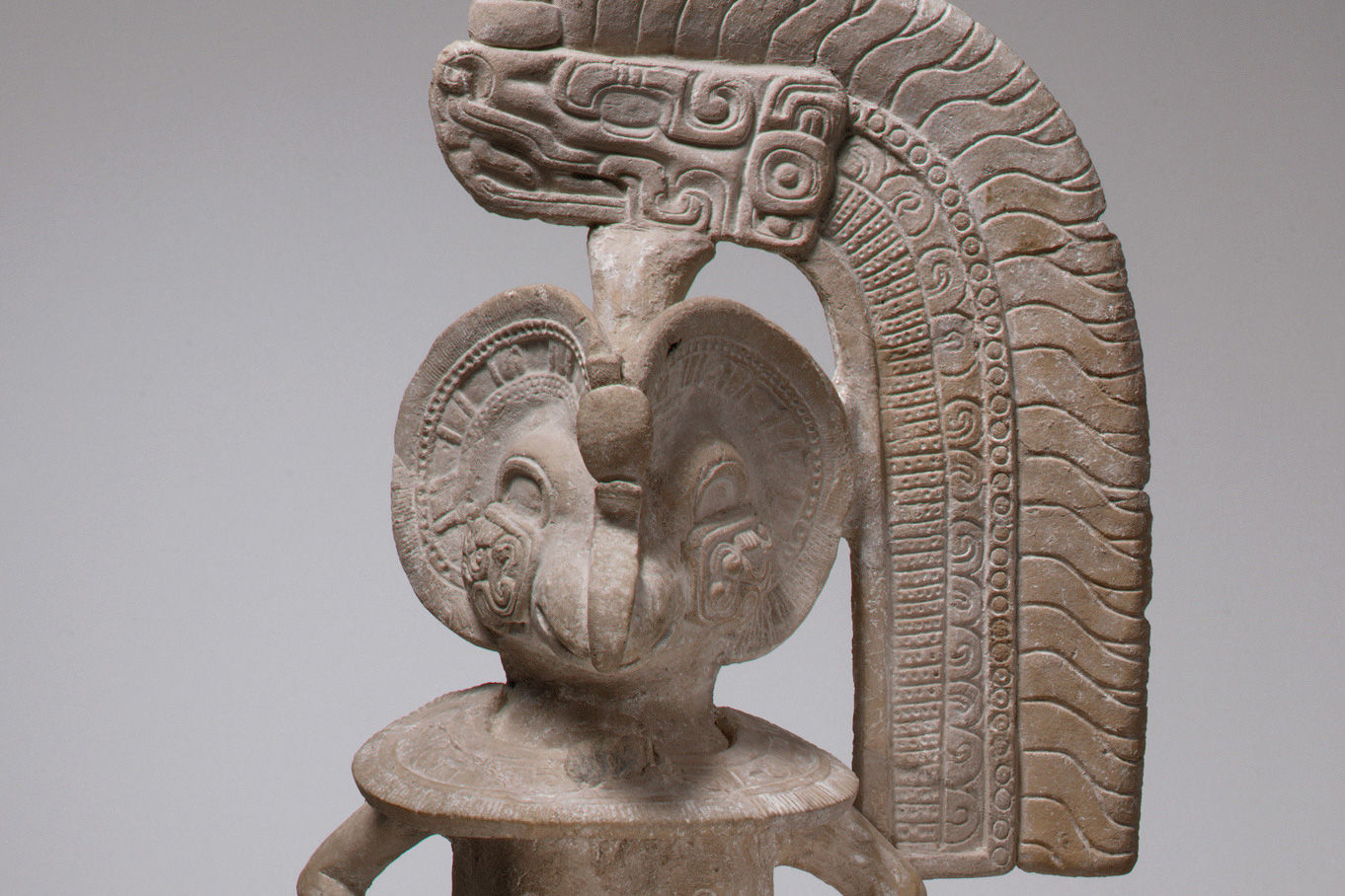 A ceramic figure wears a headdress of a feathered, horned serpent against a gray background.