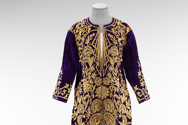 Image shows the top half of an Ottoman wedding dress known as a bindalli, made of purple velvet with gold embroidery and set against a gray background.