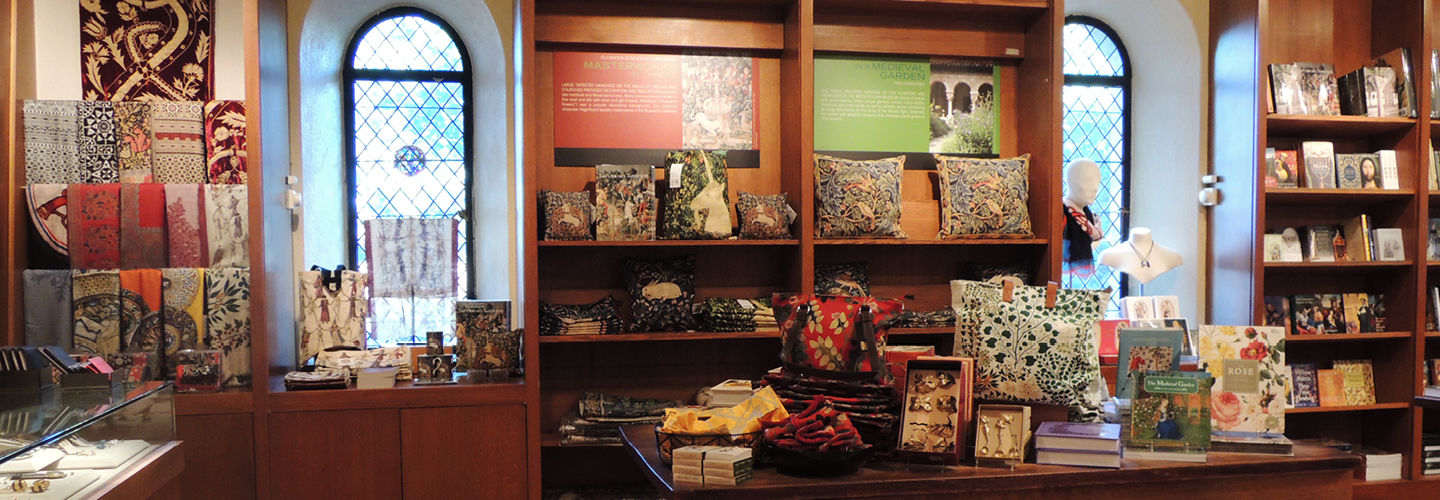 A Met Store display in The Met Cloisters showing a selection of tapestry-inspired items. Scarves and pillows with colorful intricate designs are displayed on shelves and tables.