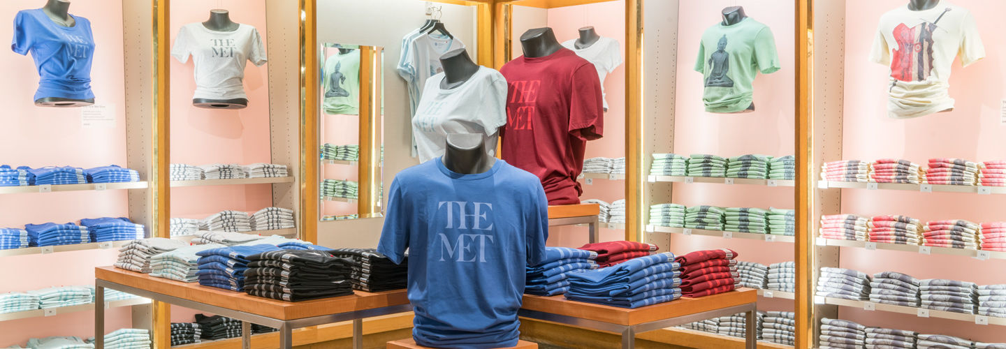 View of The Met Store showing a display of Met-branded t-shirts. The shirts are displayed on mannequin torsos without arms. The shirts are red, white, blue with text: The Met. A light green shirt with a image of buddha hangs in the background.