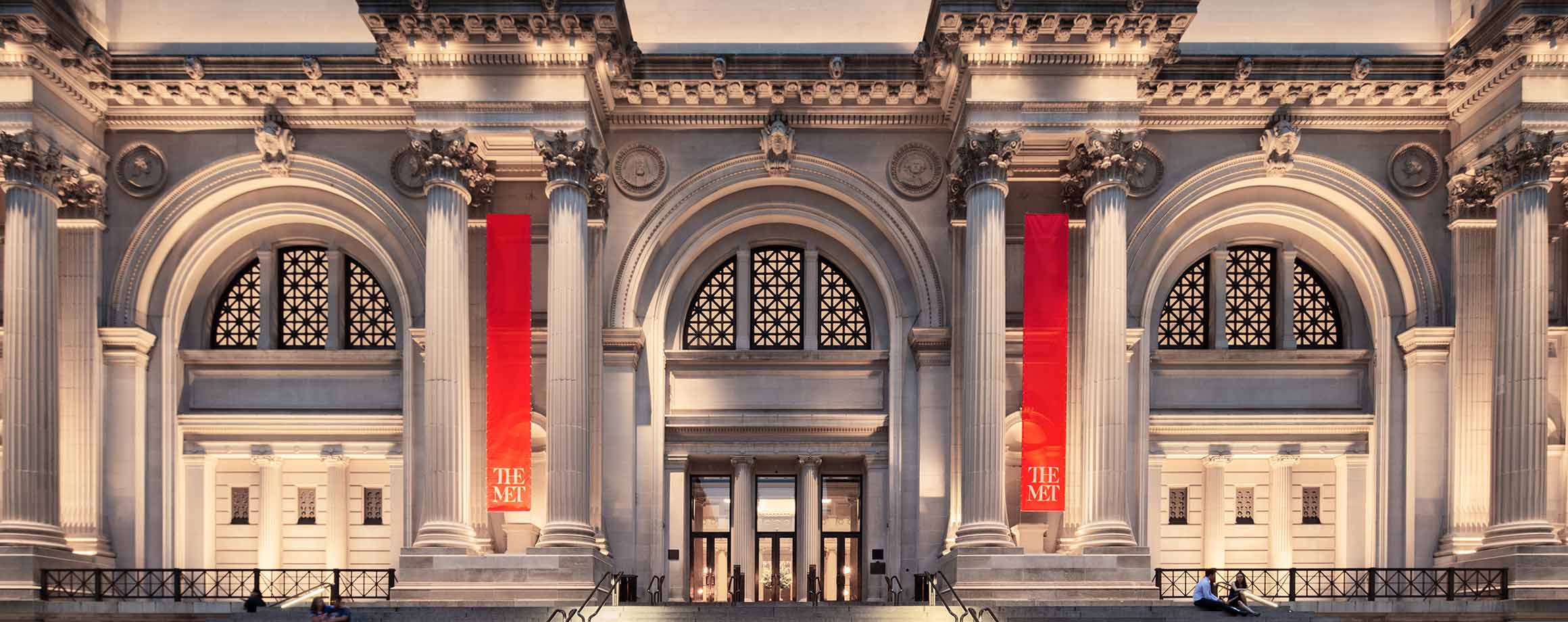 The facade of a building composed of three triumphal arches joined by huge pairs of columns with a very wide tiered staircase leading up to the center arch. Two red Met-branded banners frame the center arch.