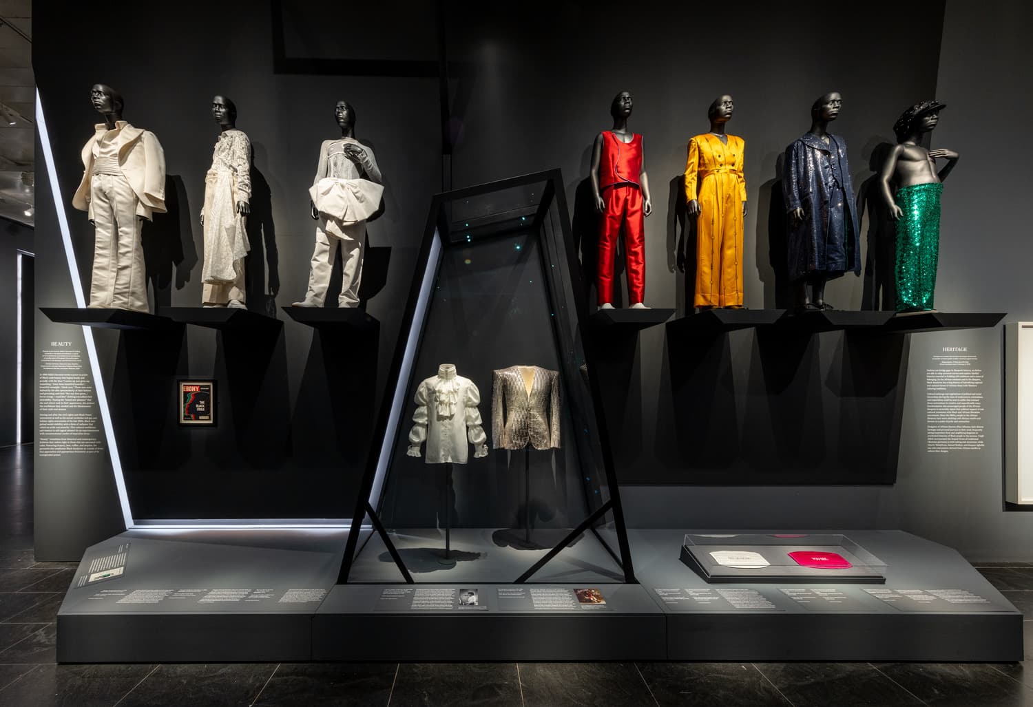 A display of nine mannequins dressed in a colorful range of fashion garments stand atop two levels of a sculptural black display. An issue of the cover of Ebony that reads “The Black Style” is framed on the left of a back wall framed by angular recessed lighting beneath a tier of mannequins. 