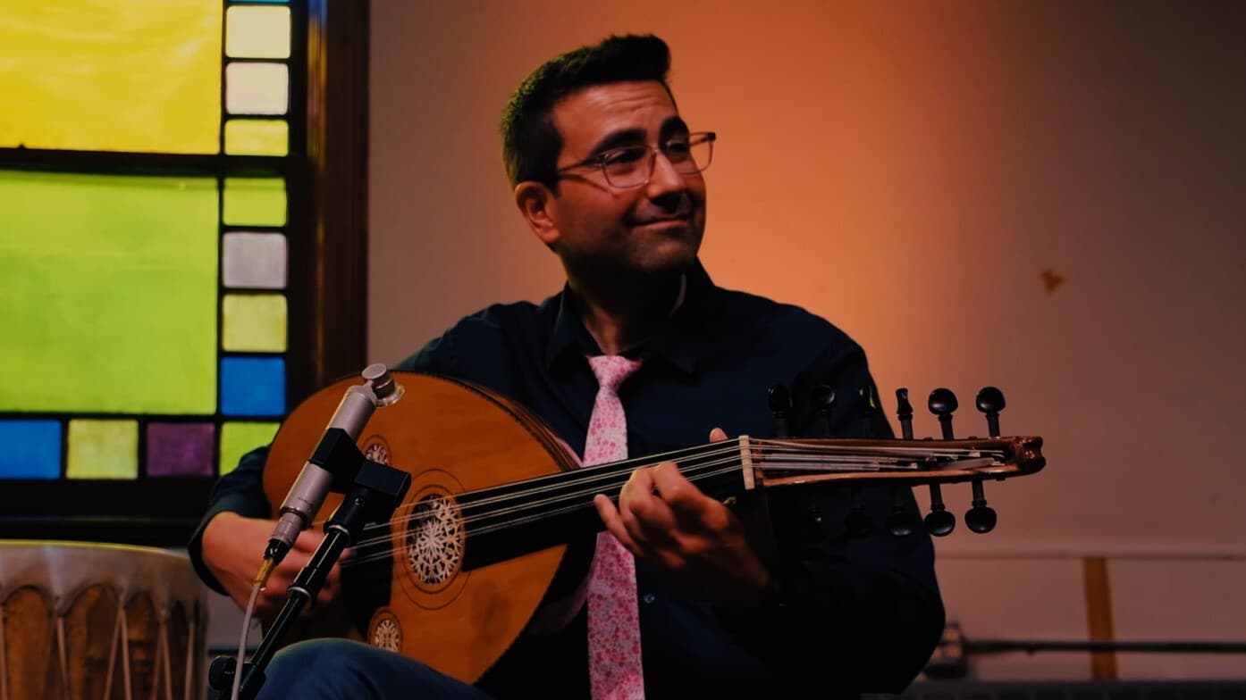 Mavrothi Kontanis, musician, holds his instrument, the oud (lute), while seated wearing a black shirt and a pink tie. 