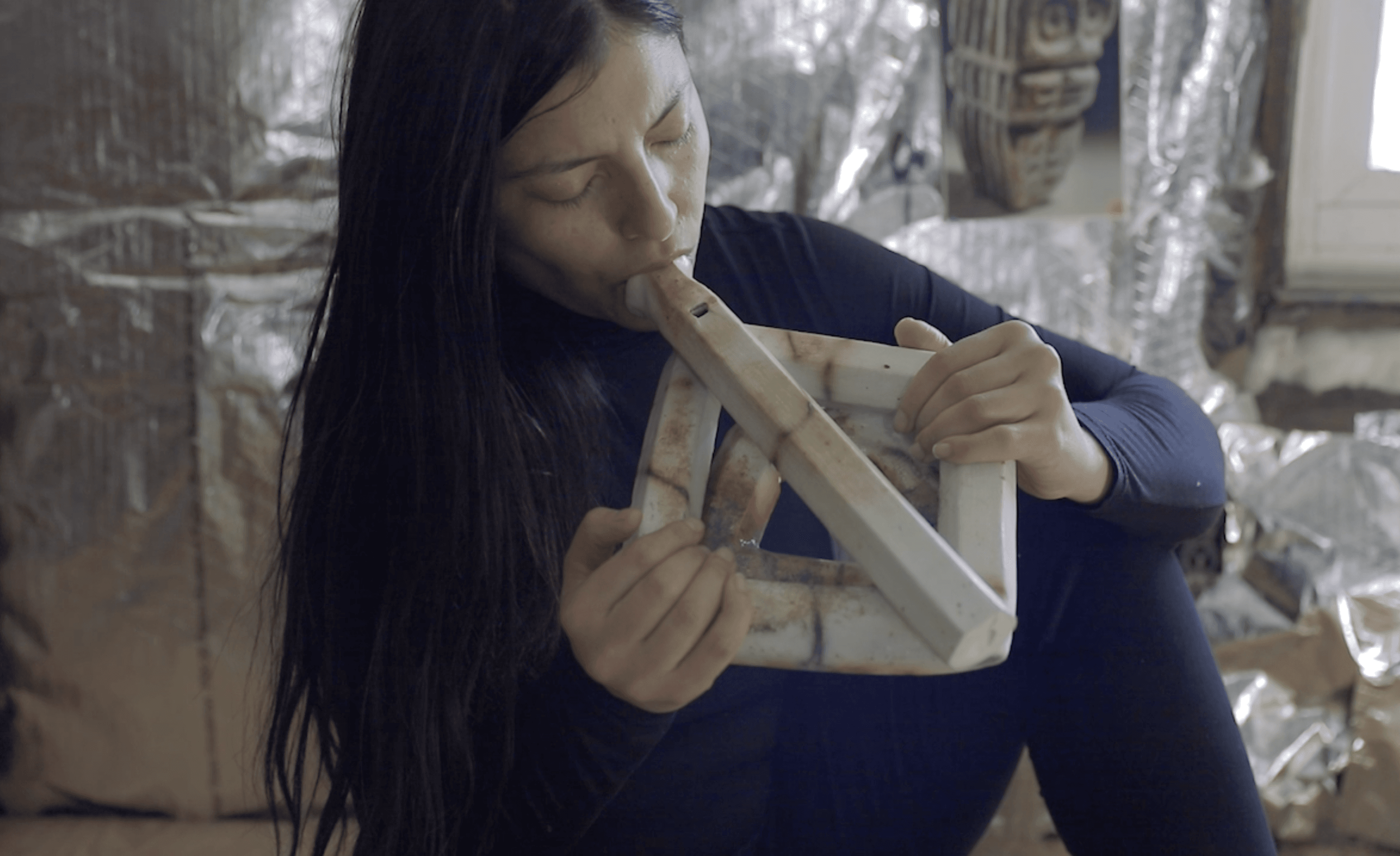 Image of artist and educator Koyoltzintli Miranda-Rivadeneira playing a large flute made of clay.