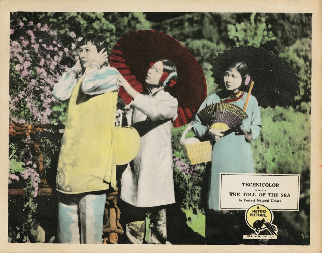 A black and white movie still printed on paper with hand coloring in pastel shades of green, yellow, blue, and pink. Three Asian women are standing in a garden, one braces the sides of her head, and two others look in her direction, both carrying parasols. A logo reading “Technicolor presents The Toll of the Sea in Perfect Natural Colors, A Metro Picture” is imprinted in the lower right corner. 