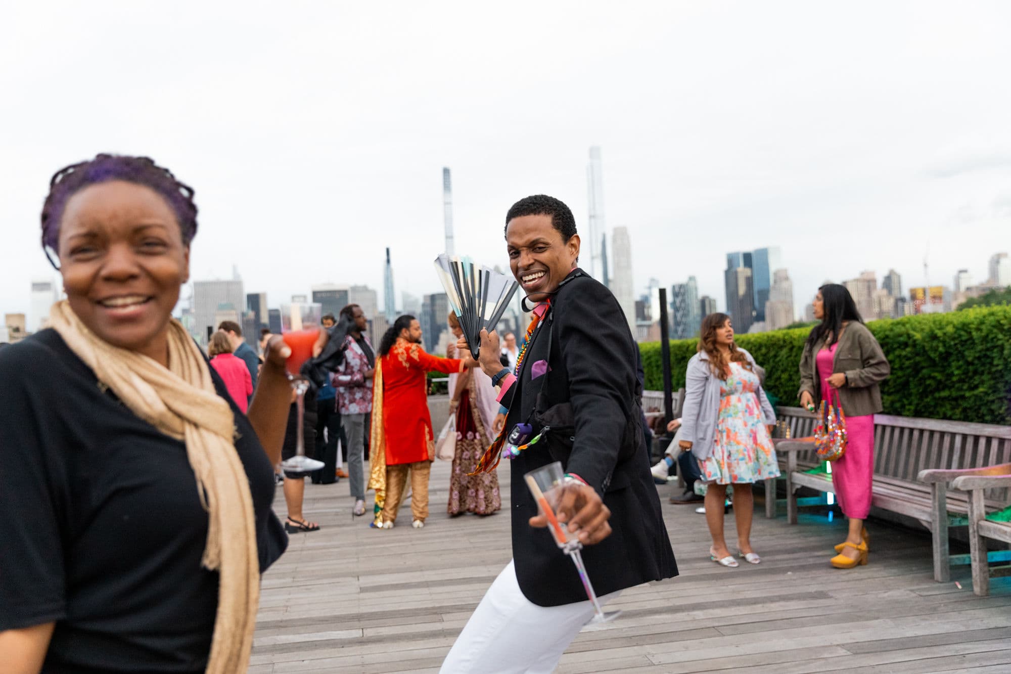 A group of people in festive attire on The Met's Cantor Roof Garden in front of the New York City skyline. Two people address the camera with laughter; one caught mid-dance. 